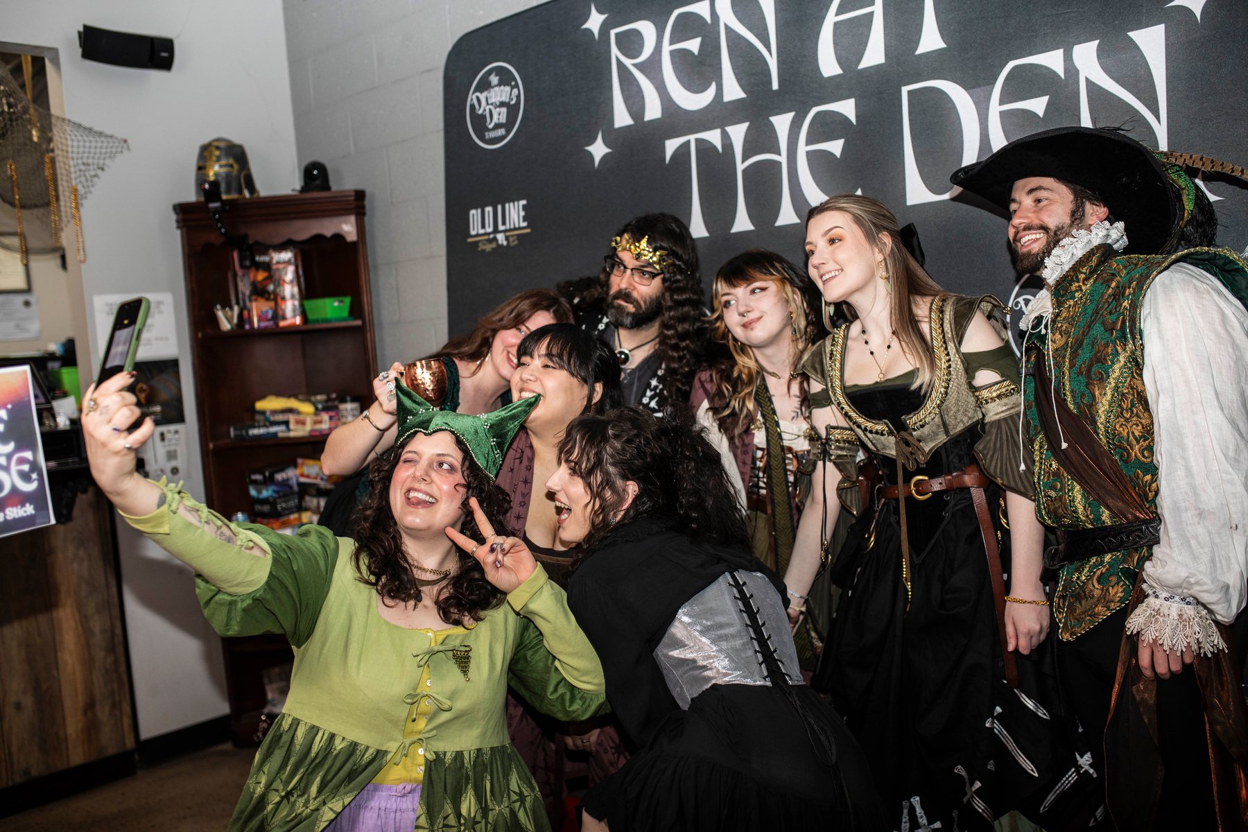 A laughing group in cloaks and corsets crowding into a selfie in front of the Ren at the Den banner