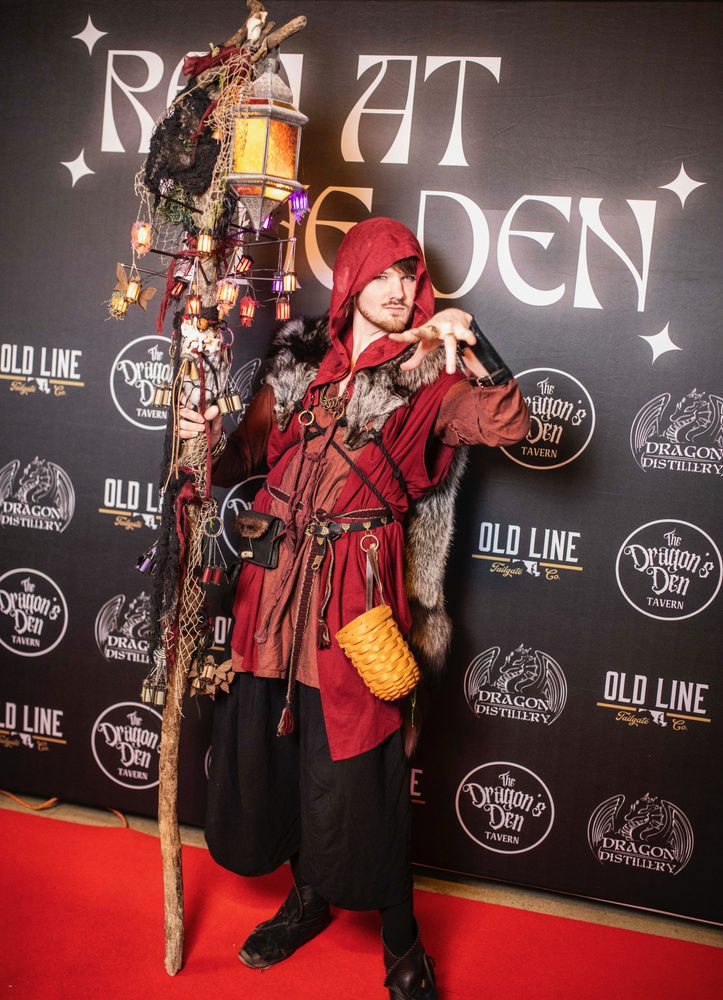 Costumed lantern-bearer in a red hooded cloak with a staff strung with glowing lanterns at the Ren at the Den photo wall